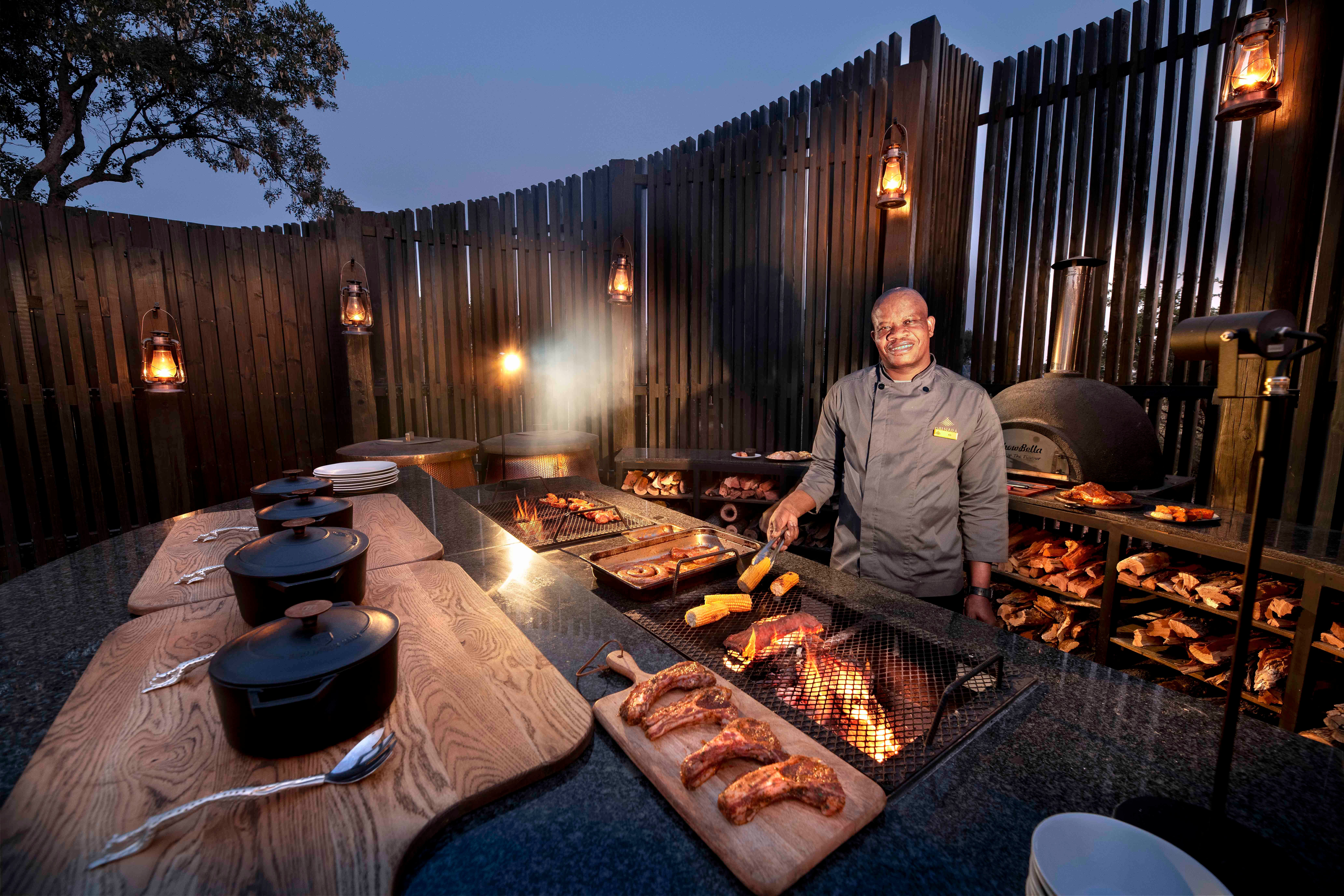 Chef Kedumetse Sefofu preparing dinner over an open fire in Daunara Safari Camp’s outdoor kitchen, surrounded by glowing lanterns and wood-fired cooking stations at dusk.