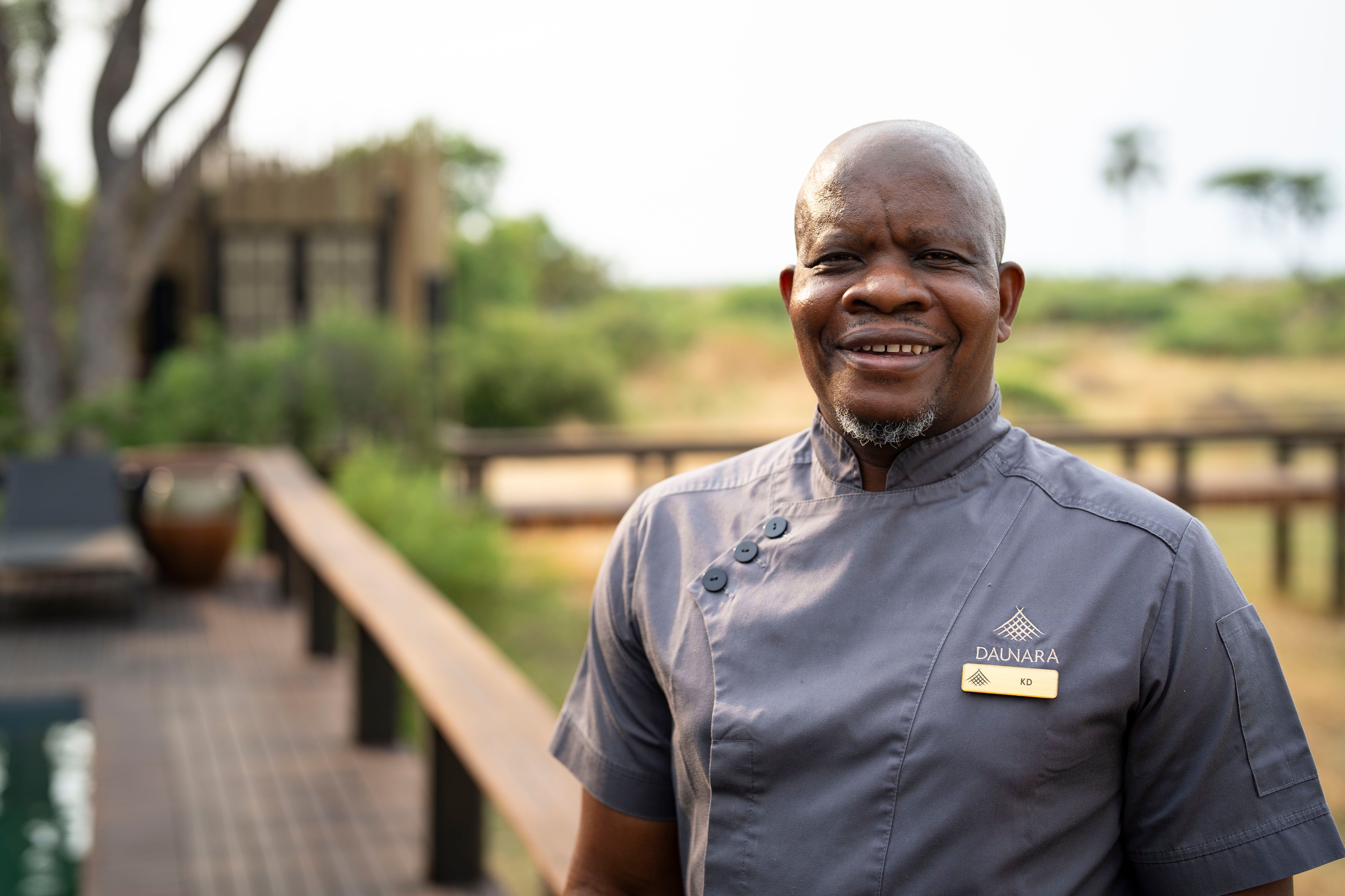 Portrait of Chef Kedumetse Sefofu wearing a Daunara Safari Camp uniform, smiling warmly on the deck with the Delta landscape softly blurred in the background.